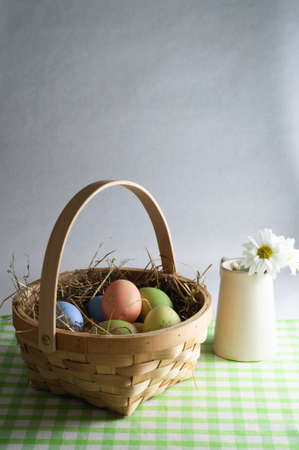 Wooden basket of decorated Easter eggs and straw on a gingham table cloth with a jug of flowers to...