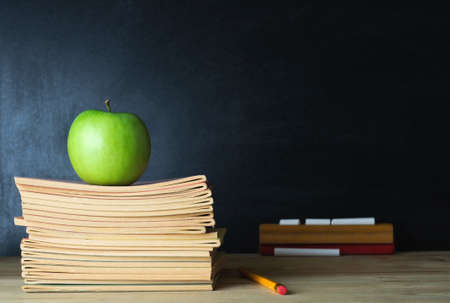 A school teacher's desk with stack of exercise books and apple in left frame. A blank blackboard in soft focus background provides copy space.の写真素材