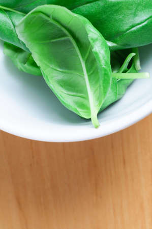 Close up (macro) of whole green basil leaves in a white china bowl, on a light wooden surface. Vertical orientation with copy space below.の写真素材