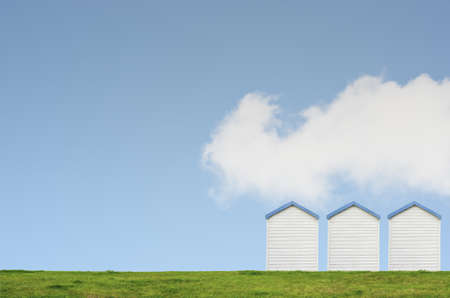 A row of three beach huts on a grassy hill against a bright blue sky with a big white fluffy cloud.  Copy space above and left.の写真素材
