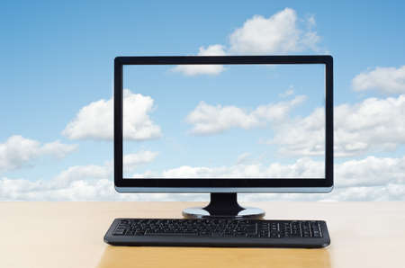 Conceptual image of cloud computing.  Computer monitor and keyboard on light wood table facing the viewer,  in front of fluffy white clouds on a blue sky, which also flow unbroken through computer screen.の写真素材