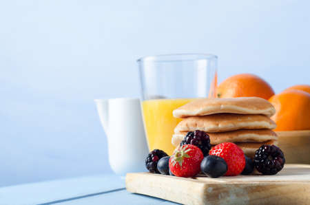 Breakfast table scene with Summer berry fruits and a stack of pancakes on an old wooden chopping board, a bowl of oranges, fresh orange juice in a glass and a white milk jug in the background.  All set on a pale blue, wood planked kitchen table with blue の写真素材
