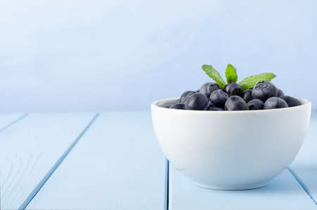 Eye level view of a white bowl of blueberries topped with a sprig of mint, on a wood planked table painted pale blue against a light blue background.の写真素材
