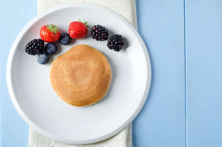 Overhead shot of a round pancake on a white plate, with Summer fruits arranged in an arc above it.  Beneath the plate is a folded hessian cloth on painted light blue wood planked table. の写真素材