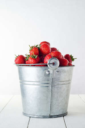 Side view of an old, pitted and scratched aluminium metal bucket filled to overflowing with harvested red strawberries on a white painted wood planked table.の写真素材