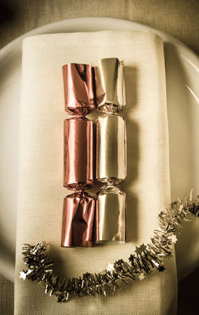 Overhead vintage style shot of red and gold shiny foil Christmas crackers resting on a napkin with a starry strip of tinsel garland beneath.  Aged photograph with vignette.の写真素材