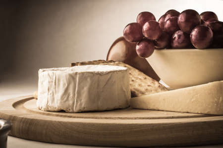 Painterly photograph of a still life arrangement of cheese, crackers, grape bowl and apple. ...