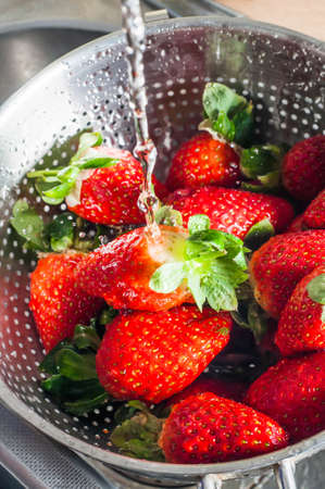 A metal sieve resting in a kitchen sink, filled with fresh strawberries and being washed under a running tap.の写真素材