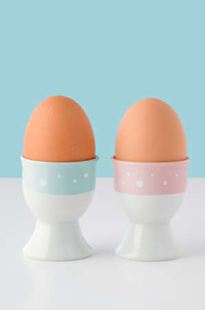 Two egg cups, decorated with pink and blue stripes with white spots and holding brown eggs on white table with light blue background.  Copy space above.の写真素材