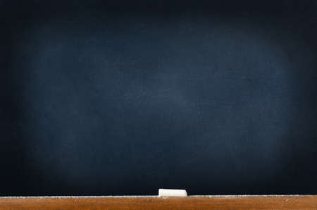 A blackboard (chalkboard) left blank to provide copy space; with a wooden ledge at the bottom, holding a piece of used chalk and sprinklings of fallen chalk dust.の写真素材