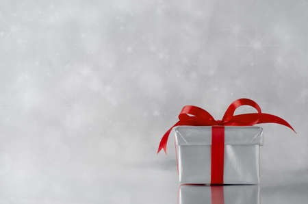 A gift box with closed lid, wrapped in metallic silver paper and tied to a bow with red satin...