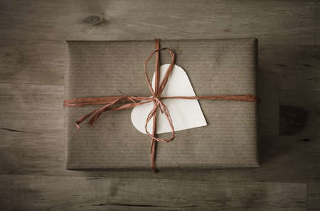 Overhead shot of a gift box with a heart shaped blank label, wrapped in brown paper with a raffia ribbon tied to a bow on wooden planked tableの写真素材