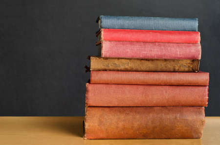 Close shot of a pile of old, shabby, well used text books stacked in a pile on a desk in front of a black chalkboard.  Copy space on left side.の写真素材