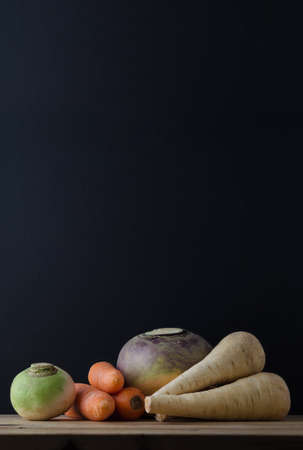 Still life arrangement of root vegetables (turnip, carrots, swede and parsnips) on a wood planked table with a chalkboard in soft focus background for copy space.の写真素材