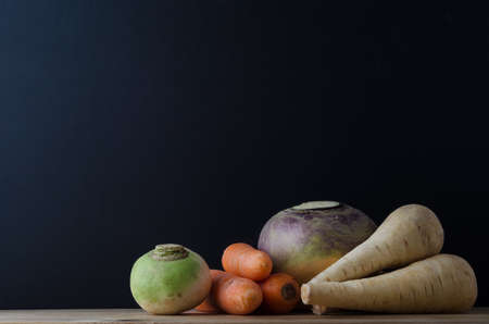 Still life arrangement of root vegetables (turnip, carrots, swede and parsnips) on a wood planked table with a blackboard in soft focus background for copy space.の写真素材