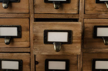 An old style wooden cabinet of library card index drawers with label holders and blank labels facing front.  One drawer in the middle is opened.の写真素材