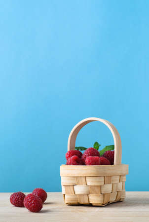 A small natural woven basket, filled with raspberries, with three grouped on the wooden surface beside it.  Blue background.の写真素材