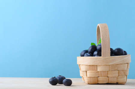 A small natural woven basket, filled with blueberries, with three grouped on the wooden surface...