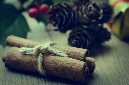 Cinnamon sticks, tied in a bundle with string on an oak table with Christmas holly, fir cones and a gold wrapped gift box in the background. Cross processed for retro or vintage effect.の写真素材