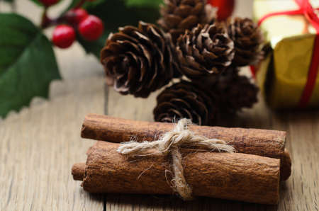 Cinnamon sticks, tied in a bundle with string on an oak table with Christmas holly, fir cones and a...