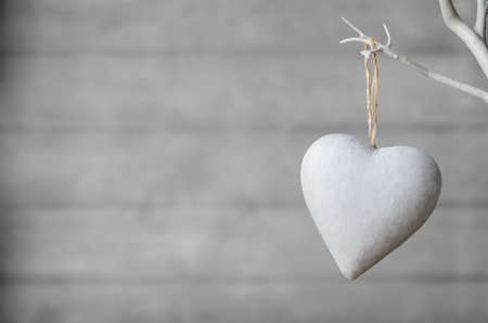 A white painted heart, hanging from branch of white artificial tree, with neutral  wood plank soft focus background.の写真素材