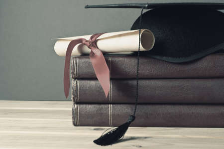 Graduation mortarboard and scroll tied with red ribbon on top of a stack of old, worn books on a light wood table.  Grey background.  Faded, washed out colours for retro or vintage appearance.