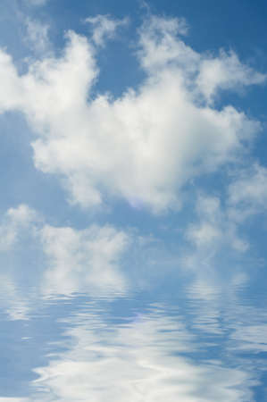Fluffy white clouds in bright blue sky, reflected in clear rippling sea or lake below horizon. The...
