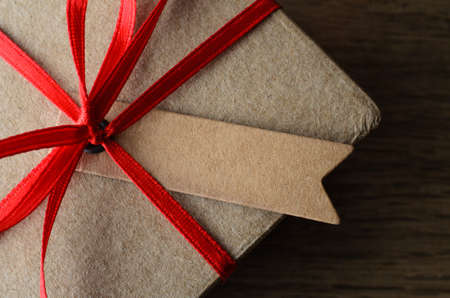 Overhead close up of a blank label tied to a plain brown  gift box with red ribbon on oak wood...