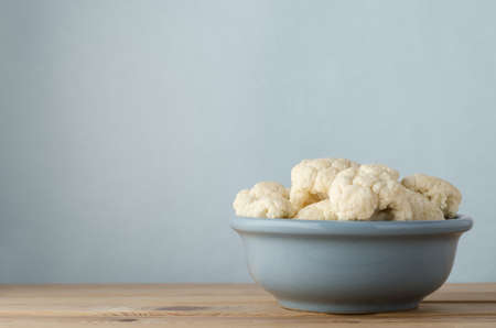 Ceramic bowl filled with raw cauliflower florets on light wood plank table against pale blue background with copy space above and to left.の写真素材