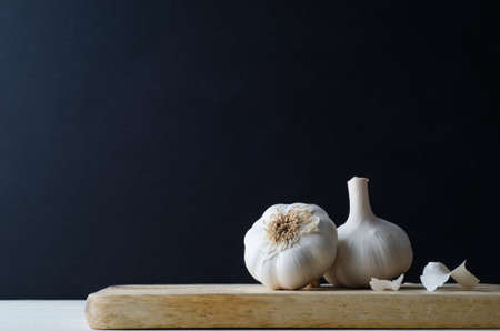 Two whole garlic bulbs on a light wood chopping board with scattered peelings. Black chalkboard background provides copy space.の写真素材