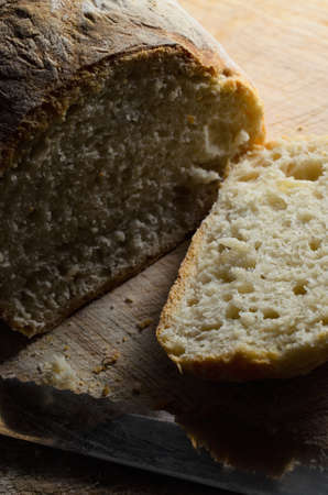 Freshly baked loaf of bread, with first slice cut and lying face up on wooden chopping board with...