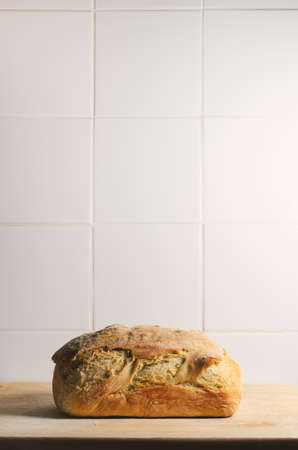 A single, whole, crusty loaf of bread on a wooden chopping board against white tiled background with...