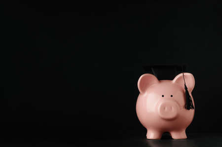 Ceramic pink piggy bank facing front at eye level wearing a black mortarboard. Blackboard background with copy space to left and above. Tuition fees concept.の写真素材