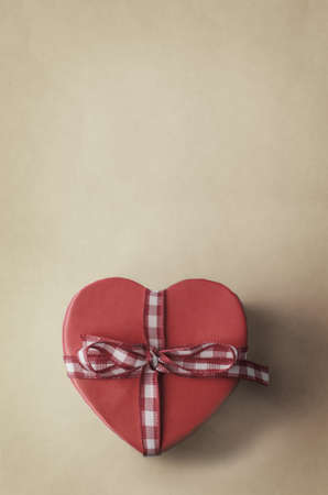 A heart shaped, undersaturated, warm pink gift box with gingham checked ribbon bow on a cream parchment style background. Copy space above.の写真素材