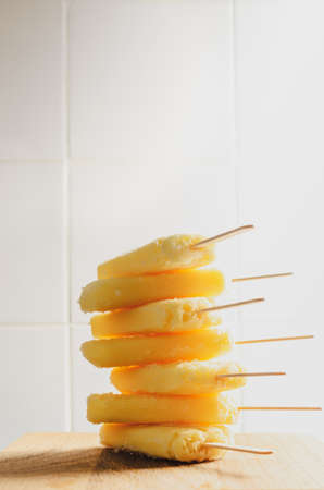Tall stack of yellow pineapple ice lollies, on a wooden table with white tiled background. Filtered...