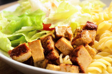 Close up of cooked tofu pieces with pasta and salad in a white bowl on light wood table. Vegetarian and vegan meal.の写真素材