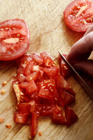 Overhead shot of hand holding knife to chop and slice juicy, red raw salad tomatoes on wooden...