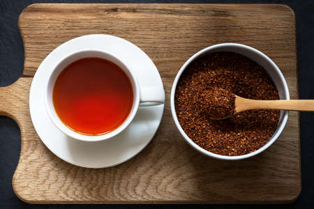 Overhead shot of a cup and saucer containing rooibos (redbush) tea with white bowl of leaves scooped in a wooden spoon. Wooden board with black slate below.の写真素材