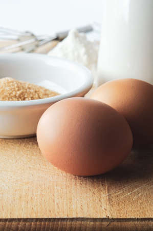 Baking preparation scene with two brown eggs in foreground, sugar, jug, flour and whisk in...