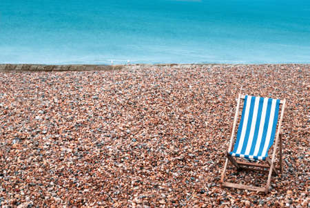 A deserted pebble beach with one empty striped deckchair in lower right foreground. Two seagulls sit on a stone groyne in the background with the sea beyond providing copy space..の写真素材