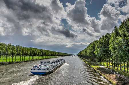 Inland barge on the long straight tree-lined Amsterdam-Rhine canal just south of Amsterdam on a day in summer with dramatic cloud formationsのeditorial素材