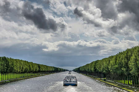 Central perspective of the long and straight Amsterdam-Rhine canal just south of Amsterdam, with inland barges and long lines of trees under a dramatic skyのeditorial素材