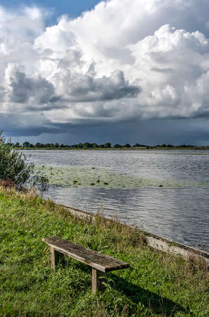 Somewhat cranky wooden bench by a lake with a view towards a spectacular cumulus cloudscape near Ankeveen, The Netherlandsの写真素材