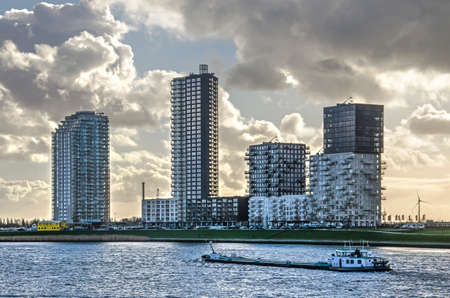 Spijkenisse, The Netherlands, January 14, 2019: a boat passes the recently created waterfront skyline on the bank of the river Oude Maas under a dramatic skyのeditorial素材