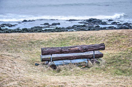 Little bench made out of tree trunks placed in the grass with a view towards a rocky beach and the...