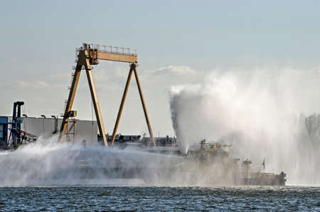 Rotterdam, The Netherlands, March 21, 2020: Port of Rotterdam fire boat spouting water on Nieuwe Maas river with a crane and harbour facilities in the backgroundのeditorial素材