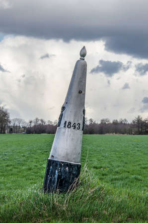 Dramatic sky over a boundery marker with inscription 1843 in a meadow on the otherwise hardly noticable border between the Netherlands and Belgium near Valkenswaardのeditorial素材