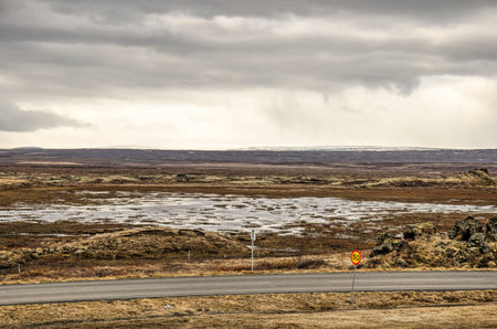 Two lane asphalt road in a desolate landscape with shallow puddles and low vegetation near lake Myvatn in Iceland under a dramatic cloudy skyの写真素材