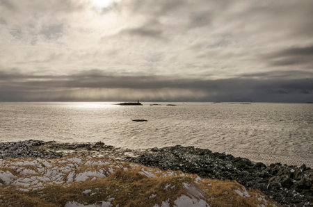 Dramatic sky over Breidafjordhur in Iceland, with a small lighthouse, a rocky beach and snow-covered...