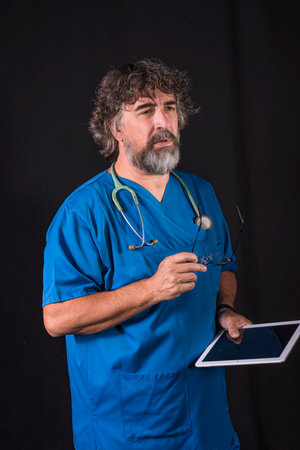 mature doctor with a beard dressed in a blue operating room suit on a black background consulting a tablet healthcare medical professionsの写真素材